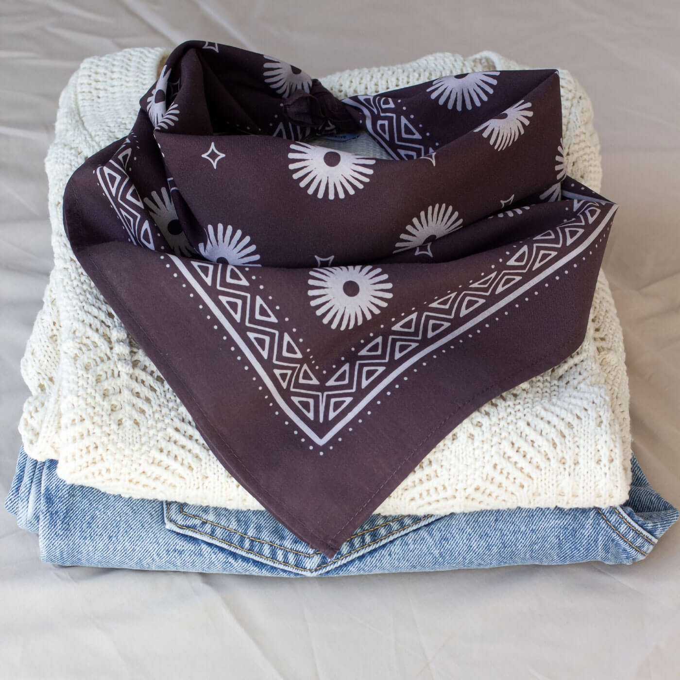 Purple bandana with white patterns on a stack of clothes including jeans and a beige sweater.