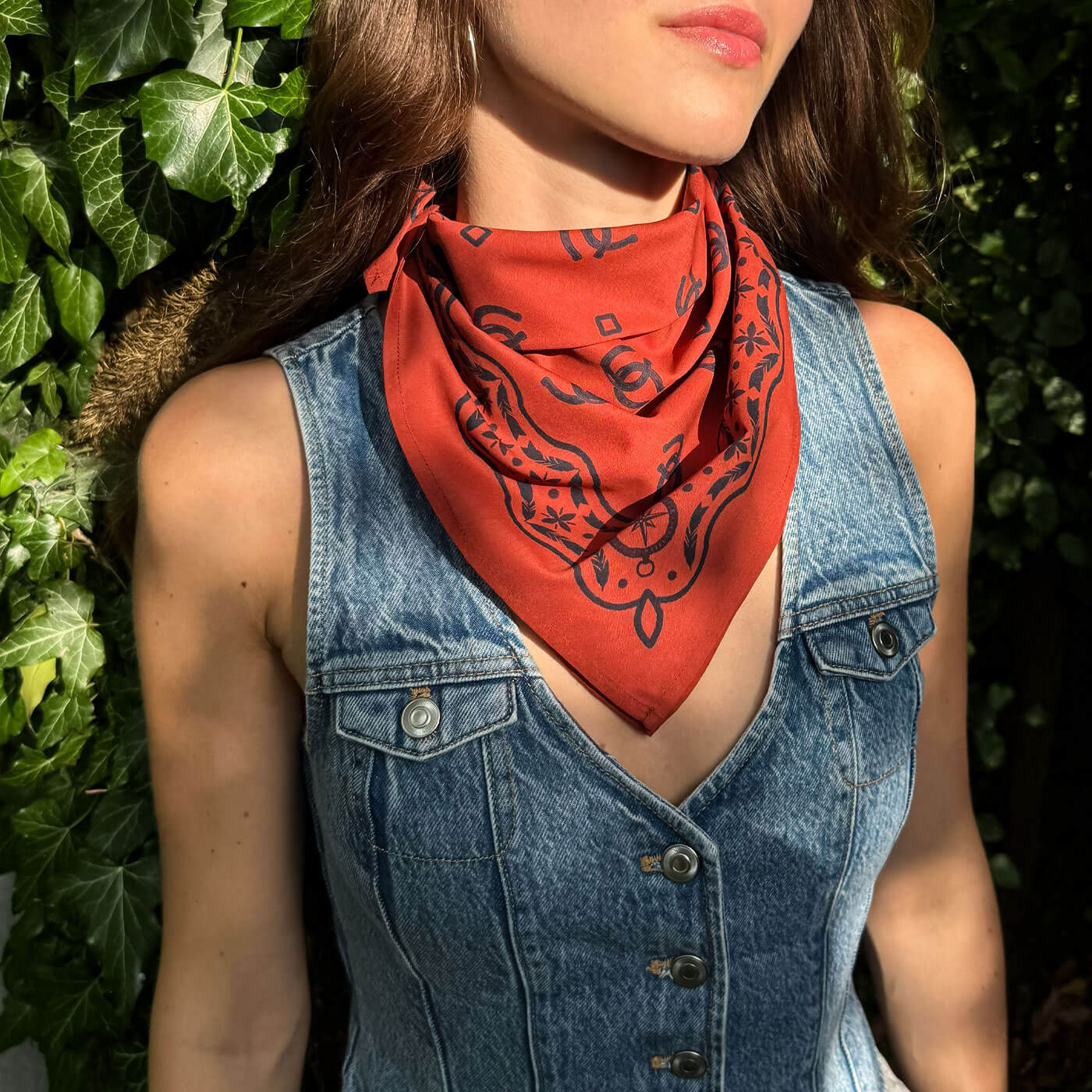 Woman wearing a red bandana with black patterns over a denim vest against a green leafy background