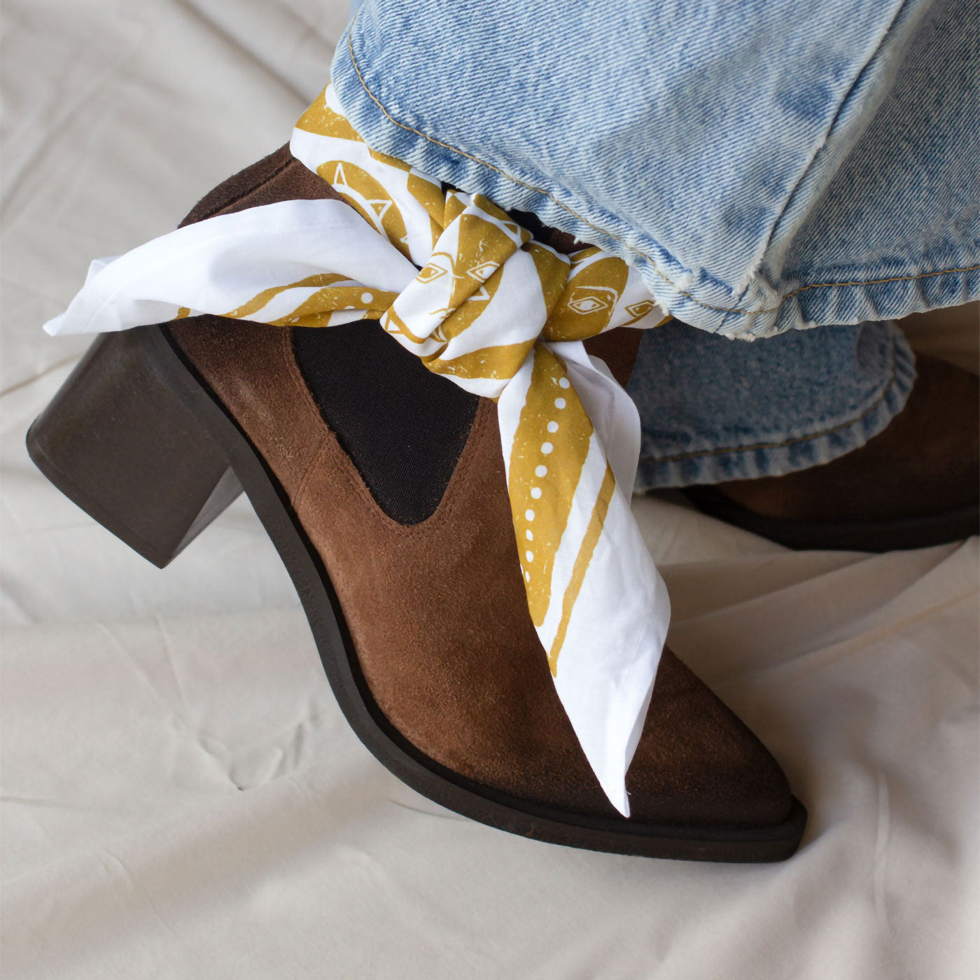 Brown high-heeled shoes with a yellow and white patterned bandana tied around them, on a light background.